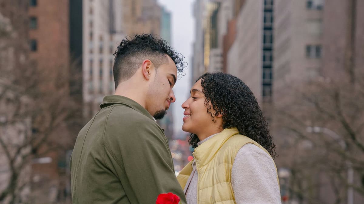Side view of positive ethnic couple with fresh red rose kissing on blurred background of urban buildings